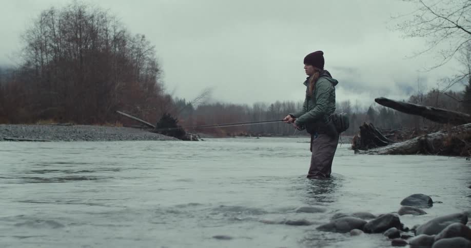 A woman standing in a river with the water up to her knees using a fishing pole to throw a fishing line