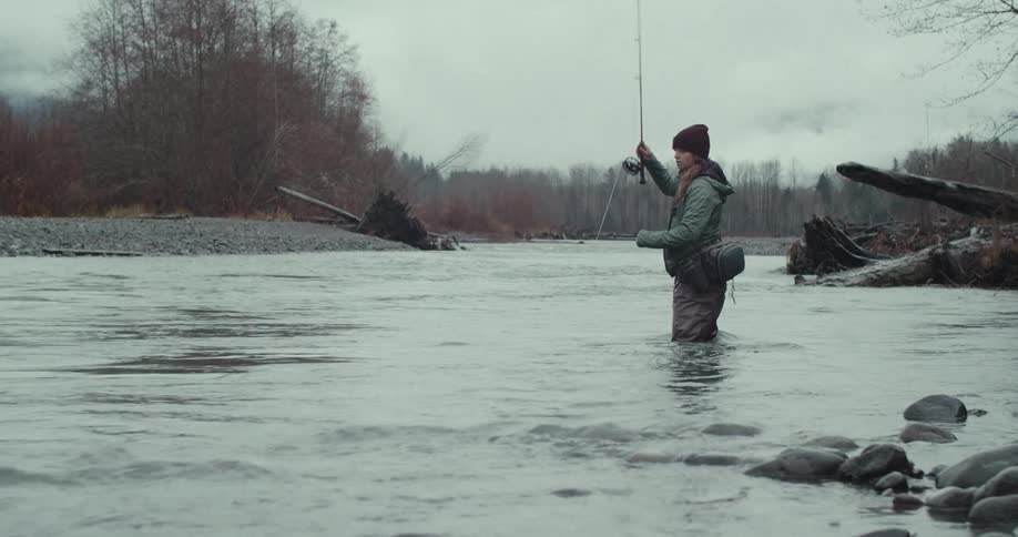 A person wearing waders and a beanie is standing in a river holding a fishing rod the person is focused on the fishing line with a riverbank and a forest of bare trees in the background the sky is grey and overcast
