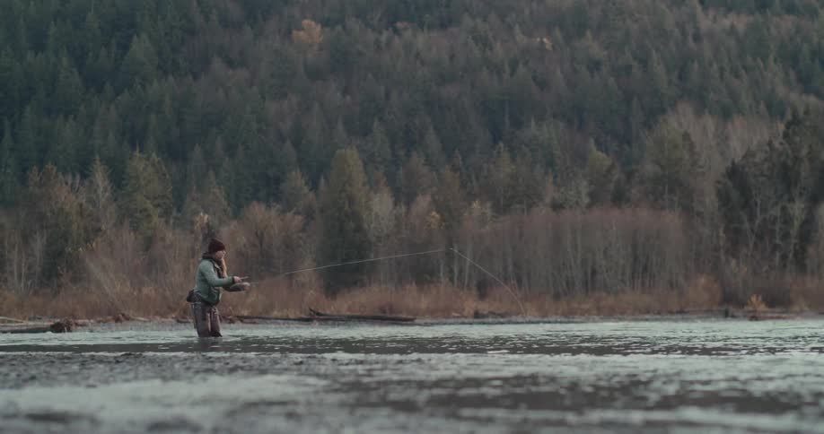 A man wearing a green jacket and waders is fly fishing alone in a shallow river with a misty treecovered mountain in the background