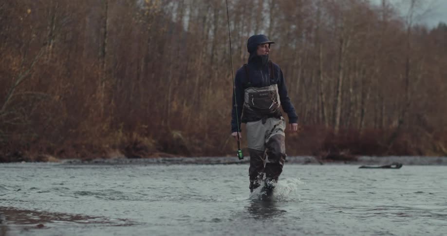 A fisherman in a hat and a dark jacket wades through a shallow river casting his line the water ripples around his legs as he patiently stands in the middle of the river surrounded by bare trees on the banks