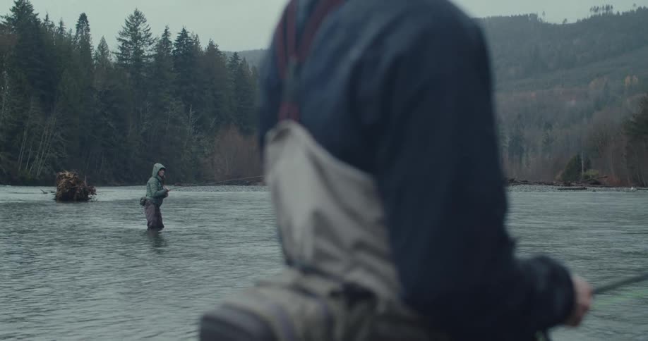 A person on a river watches while further down a woman standing up to her knees in the water tries to find a spot to throw a fishing line into the water with a fishing pole