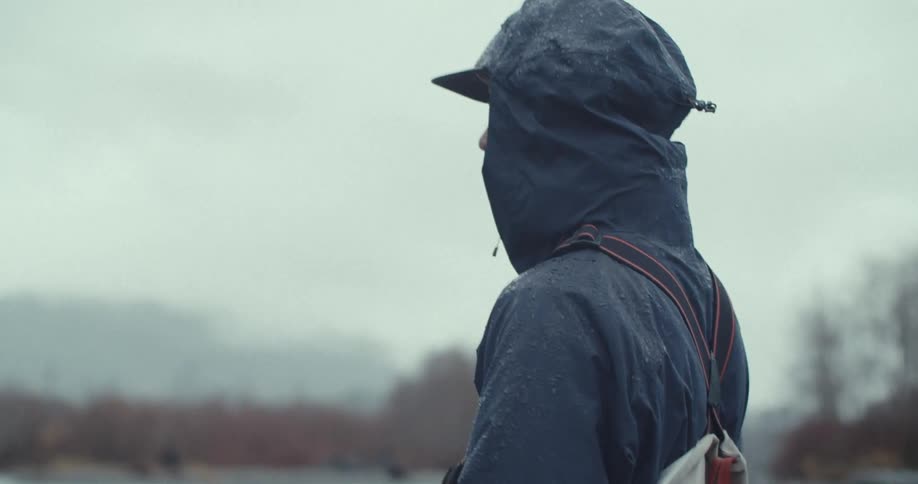 A fisherman in a hooded raincoat casts his line while standing in a river on a rainy day