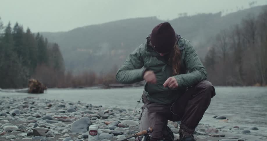 A person kneels on the rocky bank of a river and works with a fishing line with mountains and trees in the background