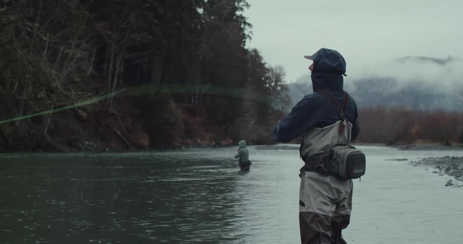 Two people stand in a river one person in the foreground casts a fishing line and another person is farther away in the water