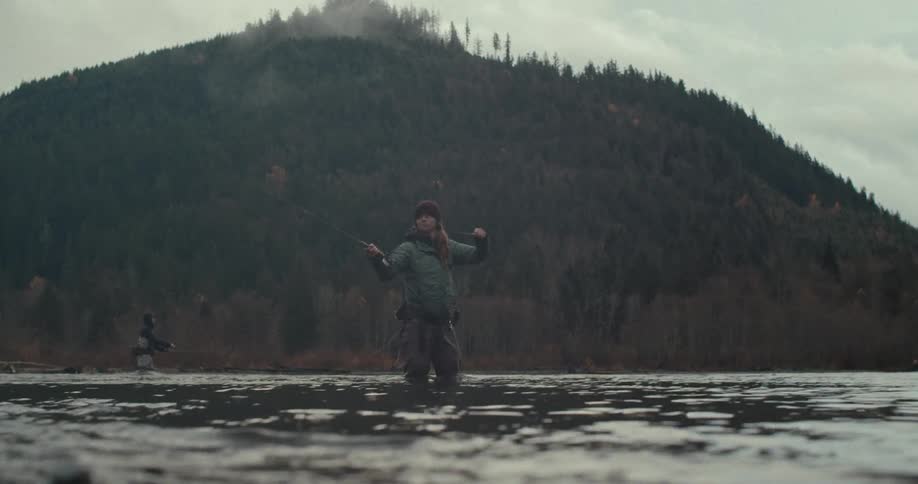 A wide shot shows a person standing waistdeep in a river fishing they are casting their line with a large forested mountain filling the background another person is visible in the distance also fishing the sky is grey and overcast