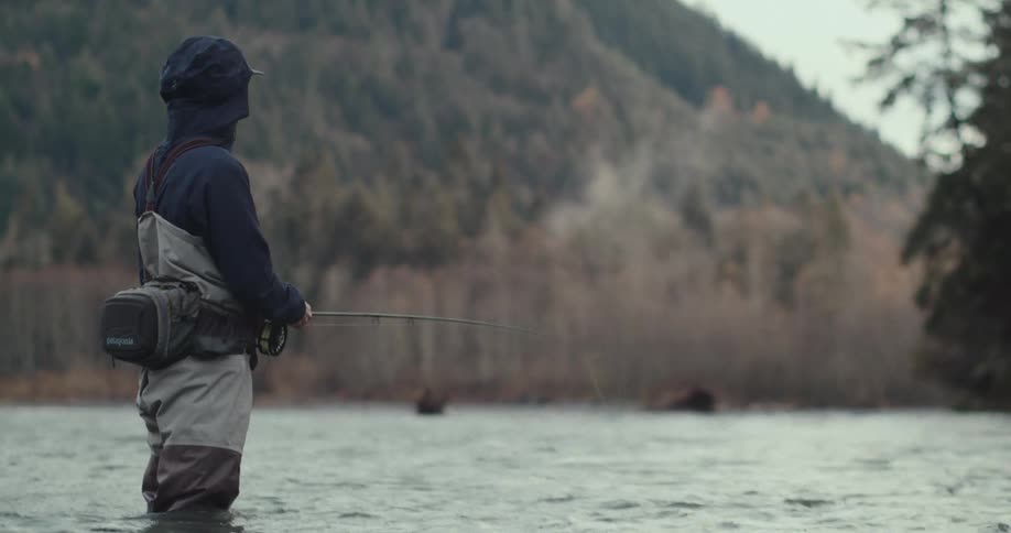 A man wearing waders and a black hooded jacket is fly fishing in a misty river with a thick treecovered mountain in the background