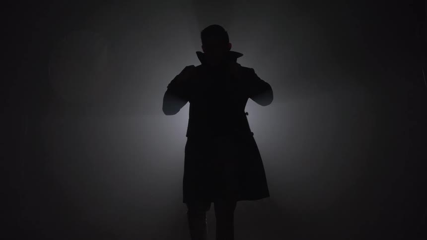 The dark silhouette of a mysterious man in a trench coat stands in a hazy backlit room adjusting his collar in a film noir style