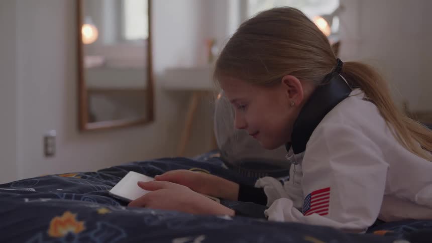 In the video a young girl wearing a white astronaut costume is lying on a bed she is holding a white tablet in her hands and looking at it intently