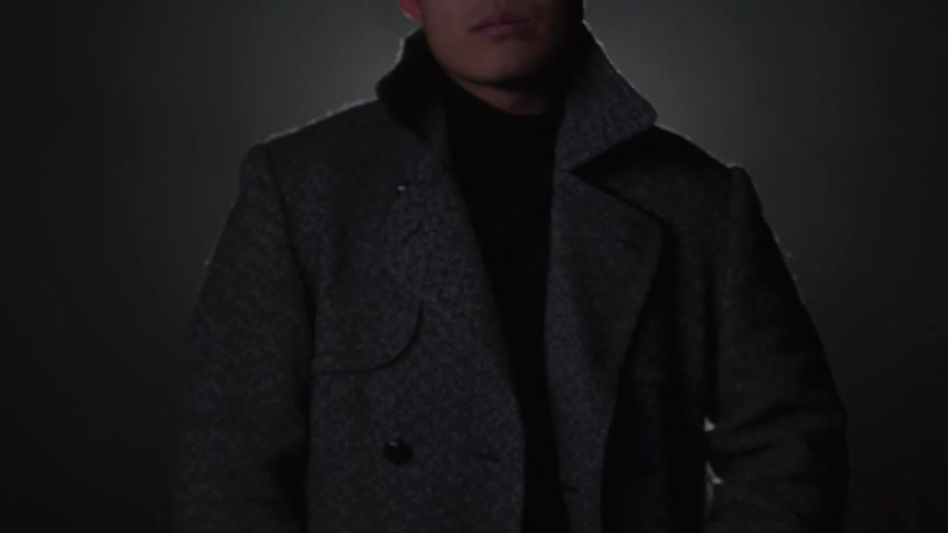 In a dark and moody shot a stylishly dressed man walks forward and stares intensely into the camera
