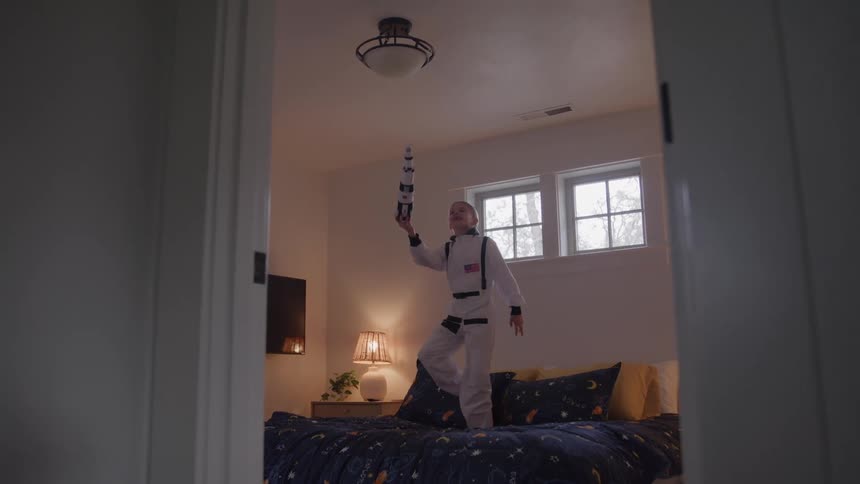A child stands on a bed wears a space suit and holds a toy while walking and jumping