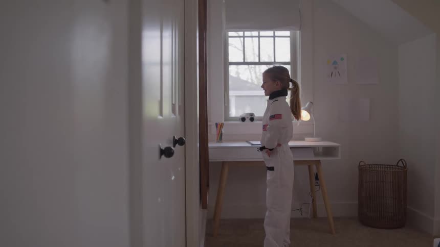 A young girl in an astronaut costume stands in her bedroom looking at her reflection in a closet door