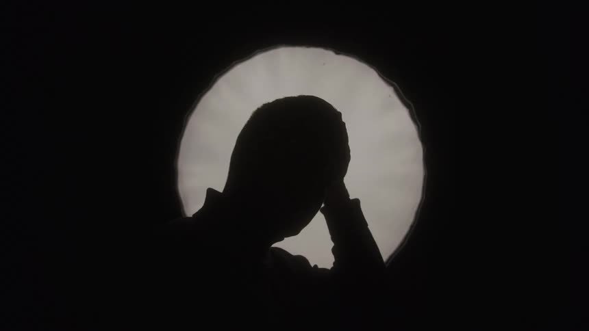 A side view of a mans silhouette is visible in a dark room through a bright ovalshaped light as he brings his hand to his head in a thoughtful manner