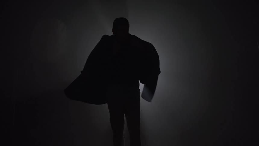 Man posing with an overcoat draped over his shoulders in a dark setting