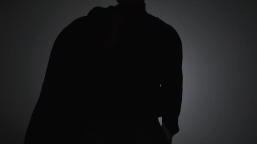 A silhouette of a person wearing a hoodie is seen in a dark room with a spotlight shining on them from the top left the person raises their right arm then slowly lowers it and eventually fades into darkness