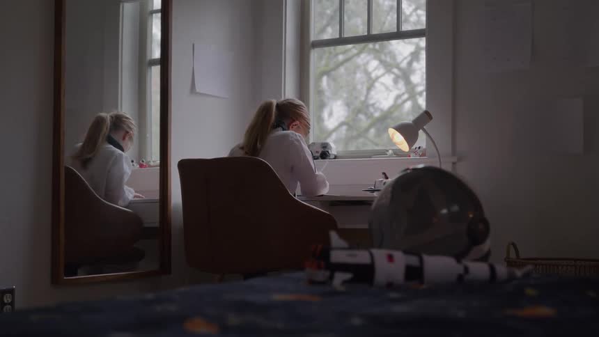 A young girl with an interest in space sits at her desk doing homework in her bedroom with a model rocket and an astronaut helmet on her bed in the foreground
