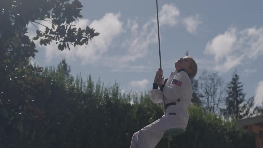 A young girl wearing a white astronaut suit is swinging on a rope swing in a grassy backyard with a bright blue sky and fluffy clouds above her