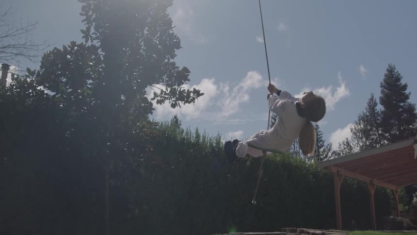 A wide shot shows a person in a white outfit swinging from a rope swing in a backyard the sun is shining behind them and trees and bushes are visible in the background under a blue sky with some clouds