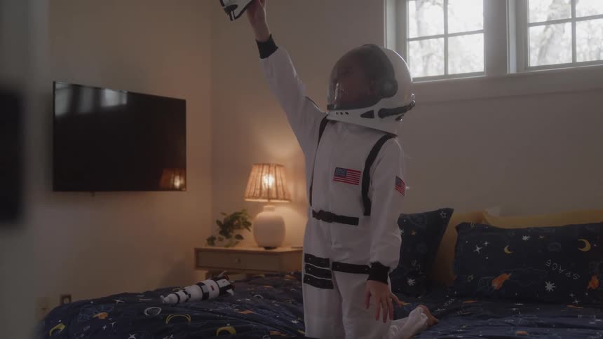 A boy wearing a white astronaut suit with an american flag on the chest is sitting on a bed with a dark blue and yellow comforter he is holding a toy rocket and raises it above his head as if he is about to launch it