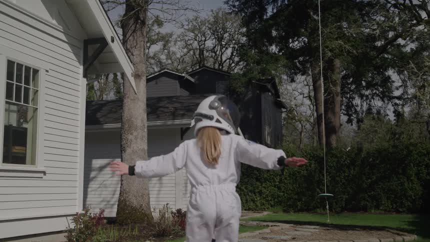 A young woman in a one piece outfit and a bike helmet runs with her arms open across a backyard