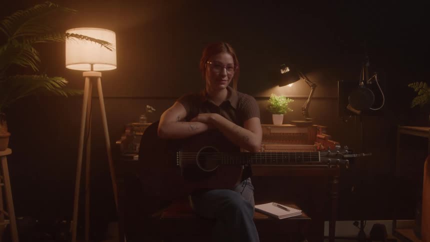 A smiling young woman sitting with her back to a piano her arms crossed over an acoustic guitar on her lap with a pen and notebook on a bench next to her