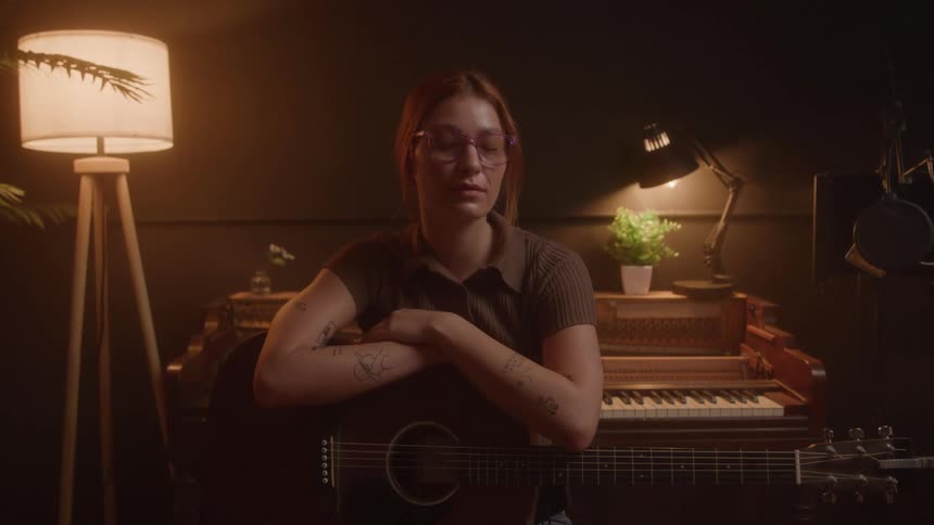 A young woman wearing a dark shirt and glasses sits in a dimly lit music studio holding a guitar and looking at the camera with a piano and two lamps in the background