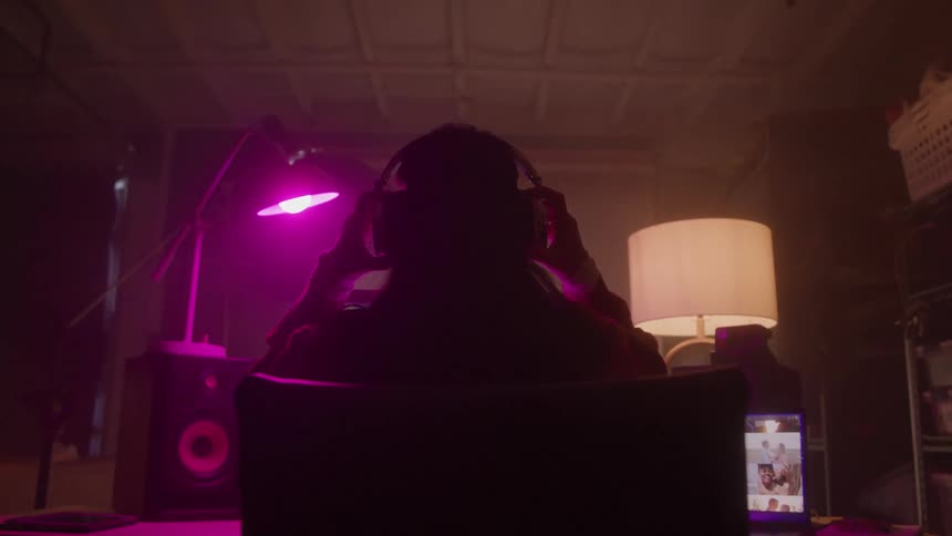 A shot from behind shows a person wearing headphones and sitting in a chair with a computer monitor in front of them the screen displays a video conference with other people in a grid the room is dimly lit with pink and purple lighting
