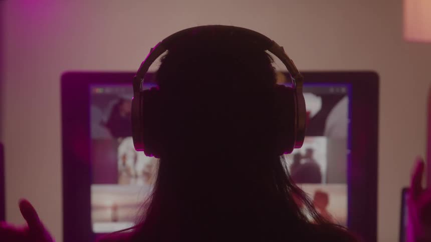 A silhouetted woman puts on headphones while on a video call on her computer in a room with pink and purple ambient light