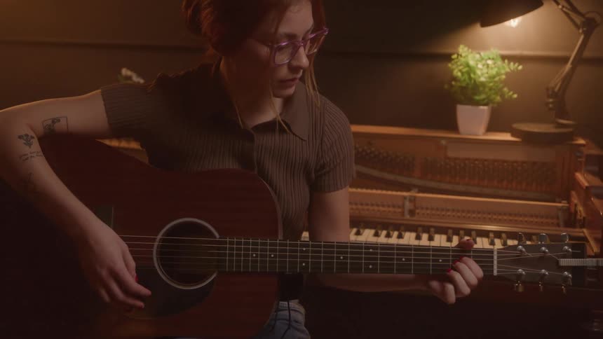 A young woman sitting with her back to a piano while playing acoustic guitar