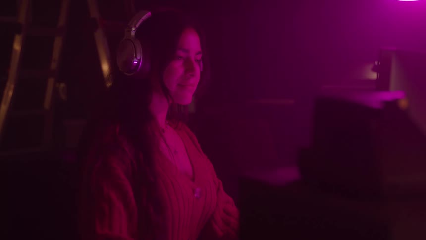 Woman in a room with pink lighting and headphones in front of a computer