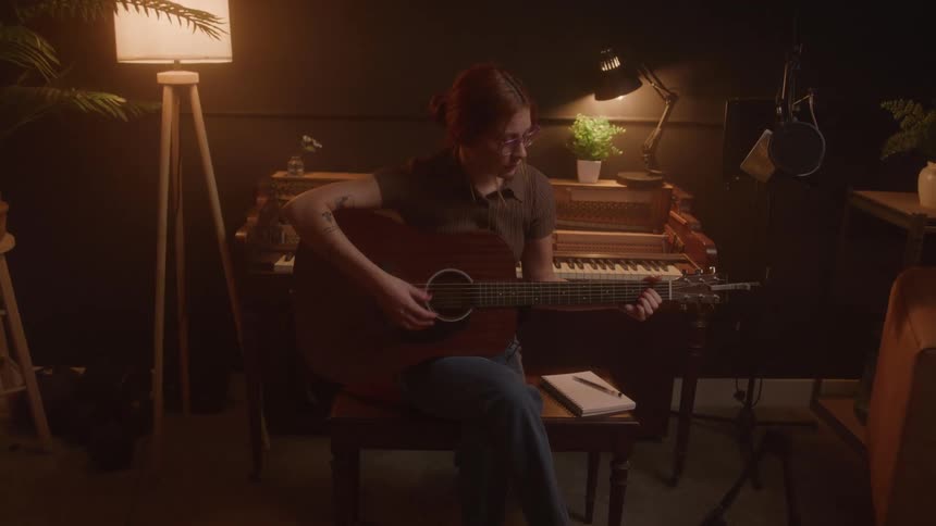 A wide shot shows a young woman sitting on a chair playing an acoustic guitar she is in a dim room with a large piano behind her and various plants and lamps surrounding her