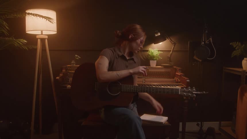 A woman with her hair in a bun and wearing a brown shirt is sitting in a dimly lit music studio playing an acoustic guitar and writing notes in a notebook