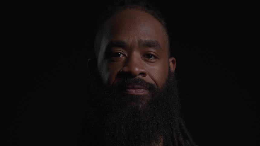 A closeup of a man with a dark beard and hair looking at the camera he has a slight smile and is standing against a dark background he appears to have a calm and thoughtful expression