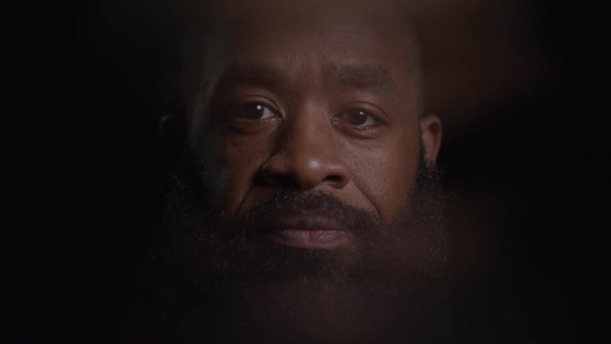 A closeup shot shows a man with a bald head and a full beard he is in a dark room and slowly raises his head to look directly at the camera he appears calm and thoughtful