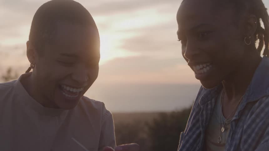 A woman in a field shows another woman a polaroid picture and they both laugh