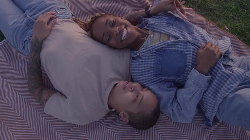 Two young female friends lying on a blanket on the lawn at sunset laughing and enjoying each others company