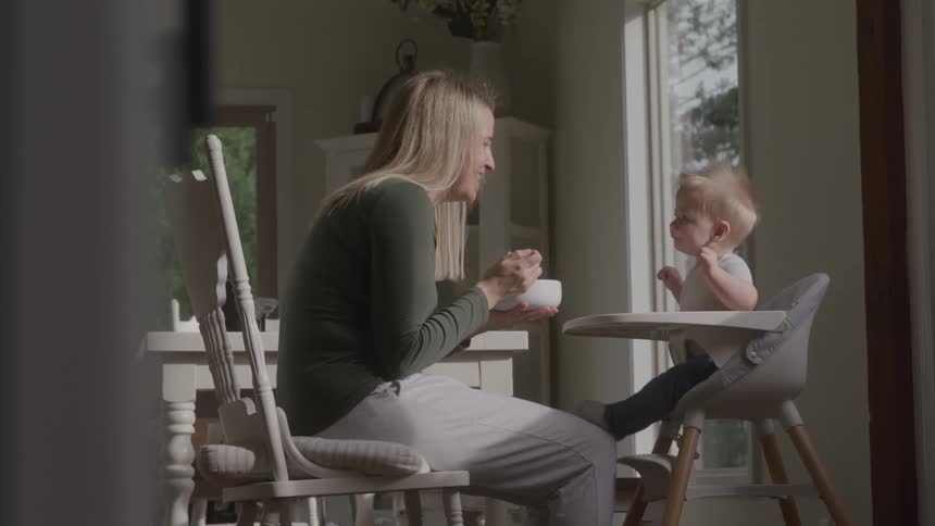 Mother and baby both laugh as the mother sitting on a wooden chair feeds the baby from a bowl with a spoon with the baby shaking his legs while sitting on a baby chair