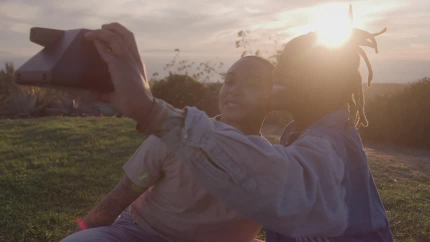 Two women sitting on a grassland at sunset taking a selfie as one kisses the other