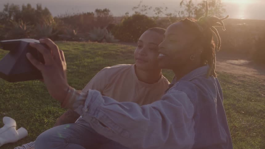 Two young people sit close together in a sunny field one of them holds a camera up and they both smile and pose for a selfie
