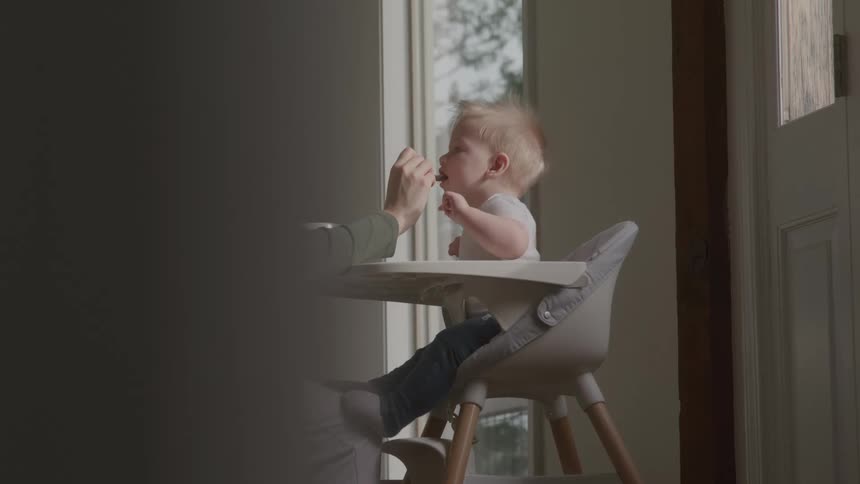 Mother feeding her toddler seated in a baby chair indoors at midday