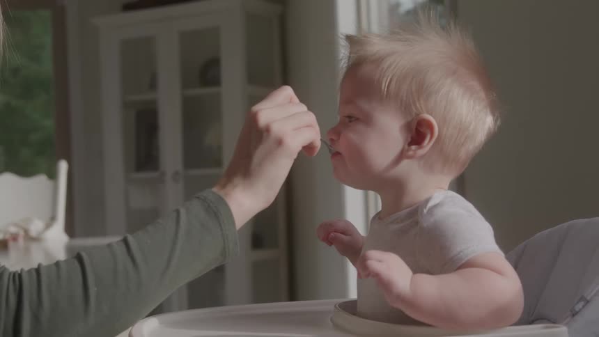 Blonde baby sitting eating from spoon given by adult person