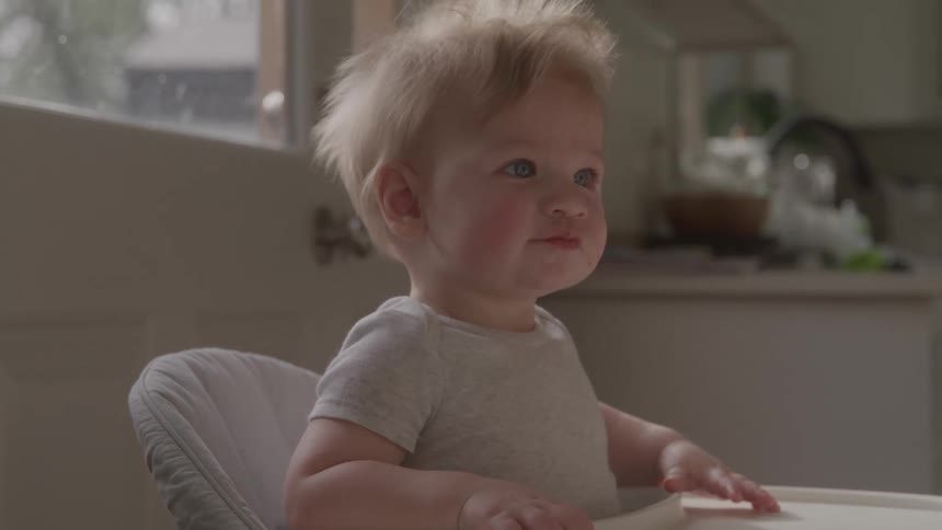 A close view shows a baby with blond hair and blue eyes sitting in a high chair looking to the side with a happy expression in a sunlit kitchen