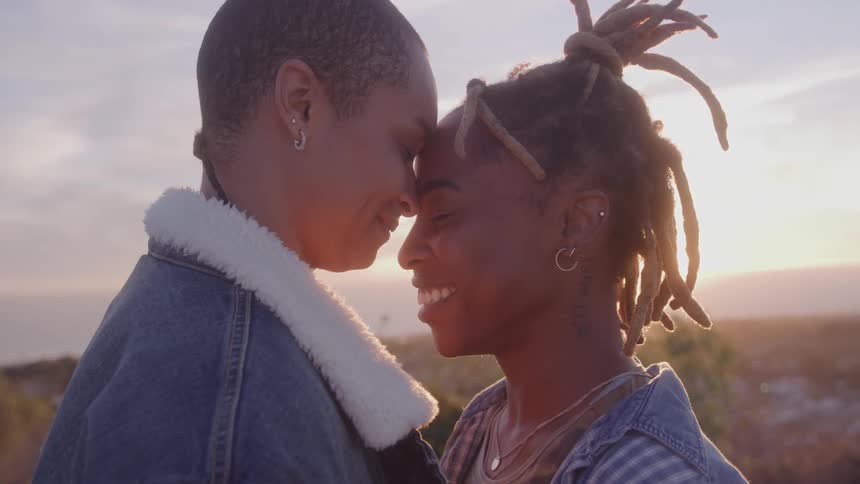 A loving couple smiles at each other while gently touching foreheads with a warm hazy sunset in the background