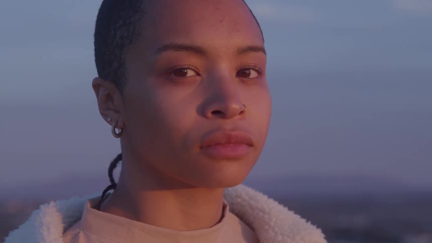 A close view shows a woman with a buzz cut and nose ring looking directly at the camera with a neutral expression as the warm light of sunset hits her face