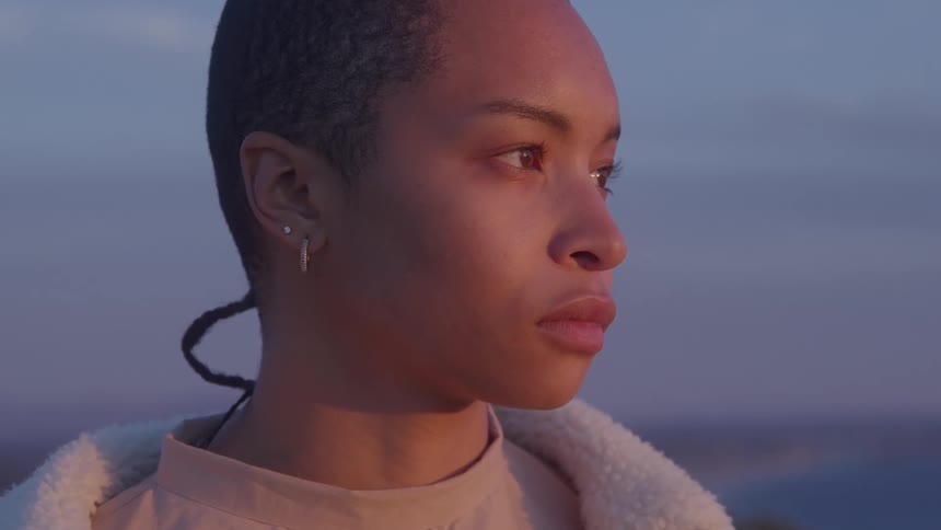 A close profile view shows a young woman with a buzz cut and nose piercing looking out at a hazy sunset with a serious expression