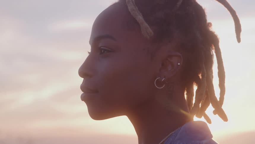 A woman stands in profile with her eyes closed her face illuminated by sunlight from behind and then she smiles