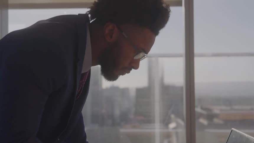 In a sunlit high rise office a young businessman in a suit and glasses stands and types on his laptop at a desk