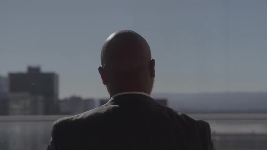 A man in a suit stands with his back to the camera and looks out a window at a city