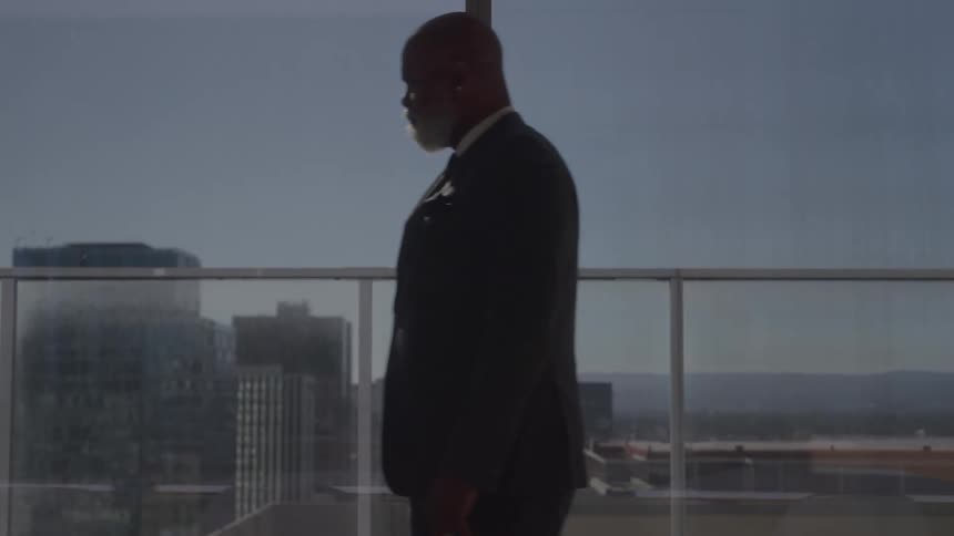 A man in a suit walks in an office setting and looks out a window at a city