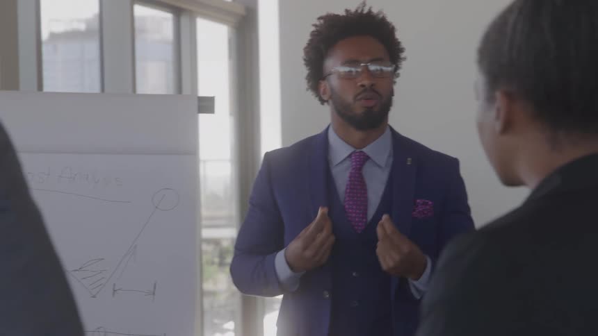 A man with glasses and a beard wearing a dark blue suit and a pink tie is standing in front of a white board and talking to another person in a sunlit office
