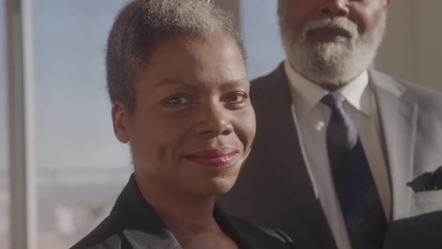 Black woman in a black suit standing in front of a black man with a grey beard wearing a grey suit inside an office building at midday with a soft smile on her face